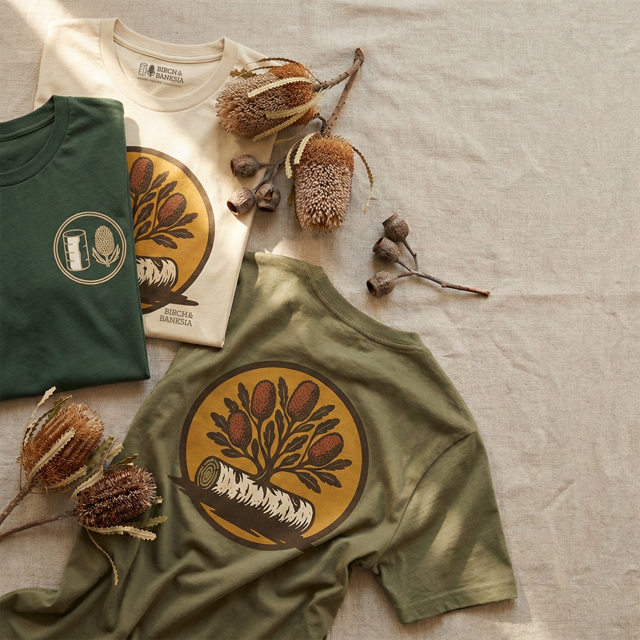 Three t-shirts with nature-themed designs and dried plants on a textured surface